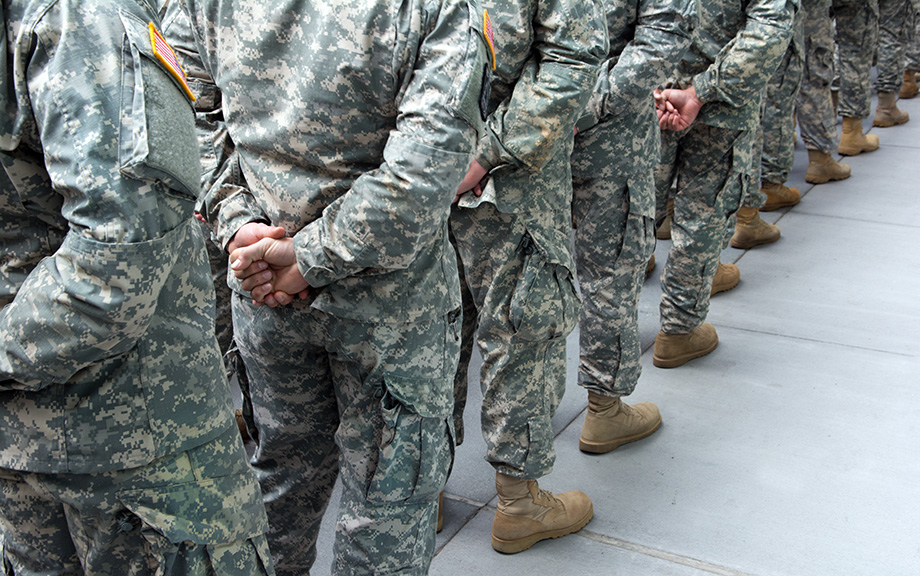 Photo: people in army fatigues lined up single file looking at their backs with the hands clasped behind them.
